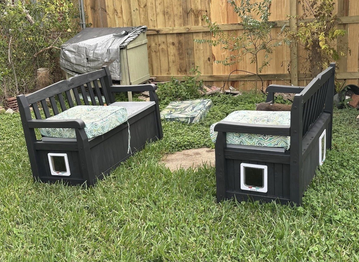 Two dark gray garden benches whose bases are enclosed shelters with small cat-flap doors, topped with patterned cushions
